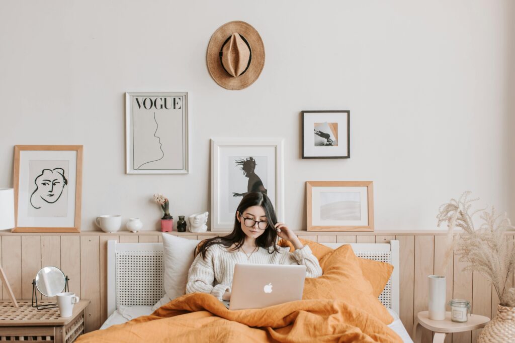 Young woman in cozy bedroom using a laptop, surrounded by stylish decor and art.