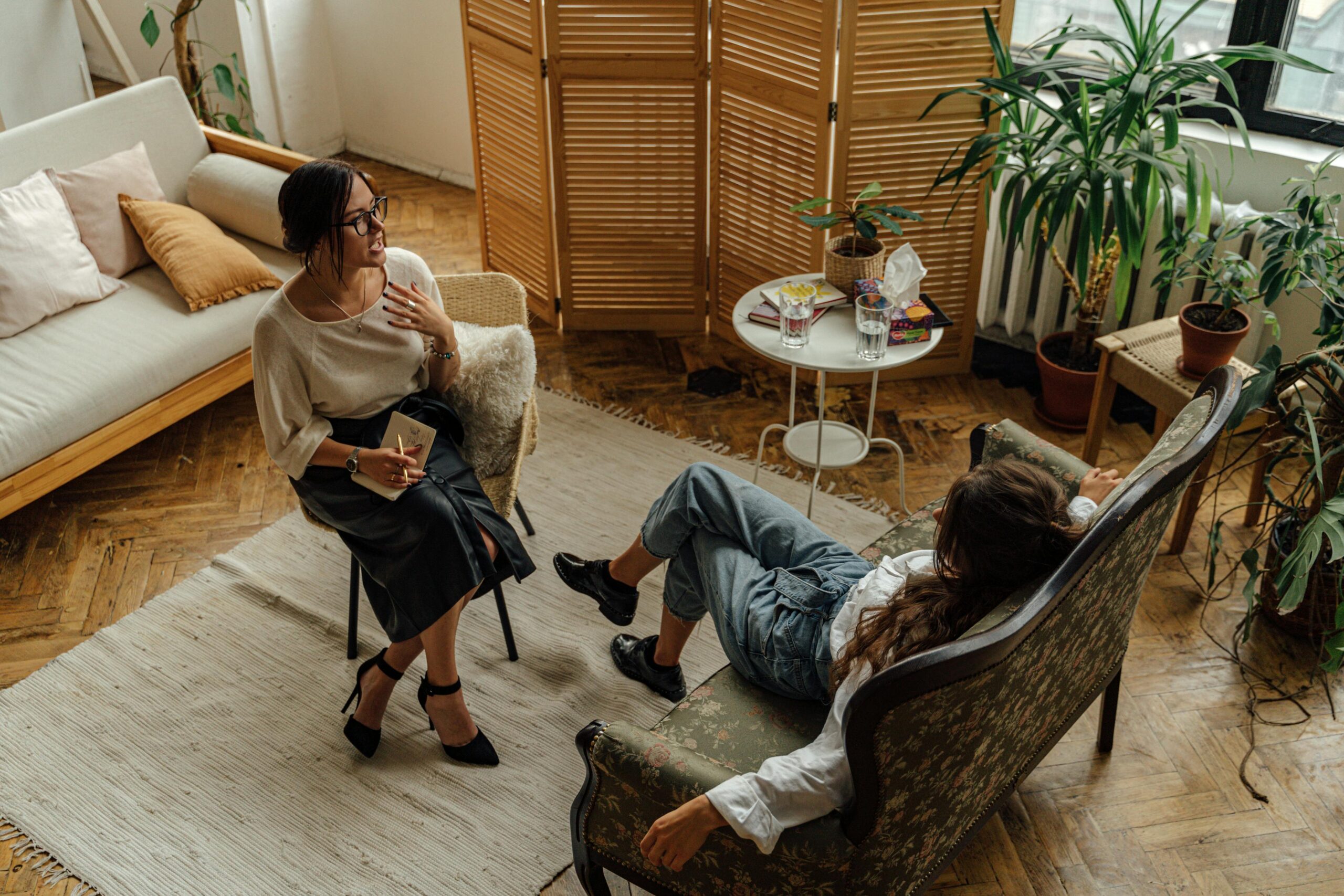 Guilt-Tripping: When Blame Becomes a Defense Mechanism Two women engaged in a psychotherapy session in a warm, inviting interior with plants and natural lighting.