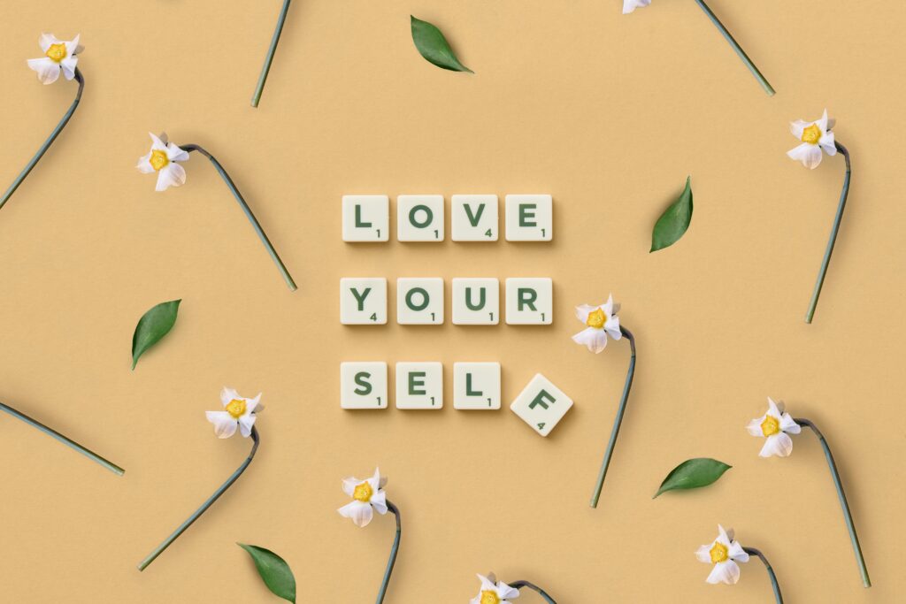 pexels photo 6005373 6005373 Scrabble tiles spelling 'Love Yourself' surrounded by flowers and leaves on a flat surface.